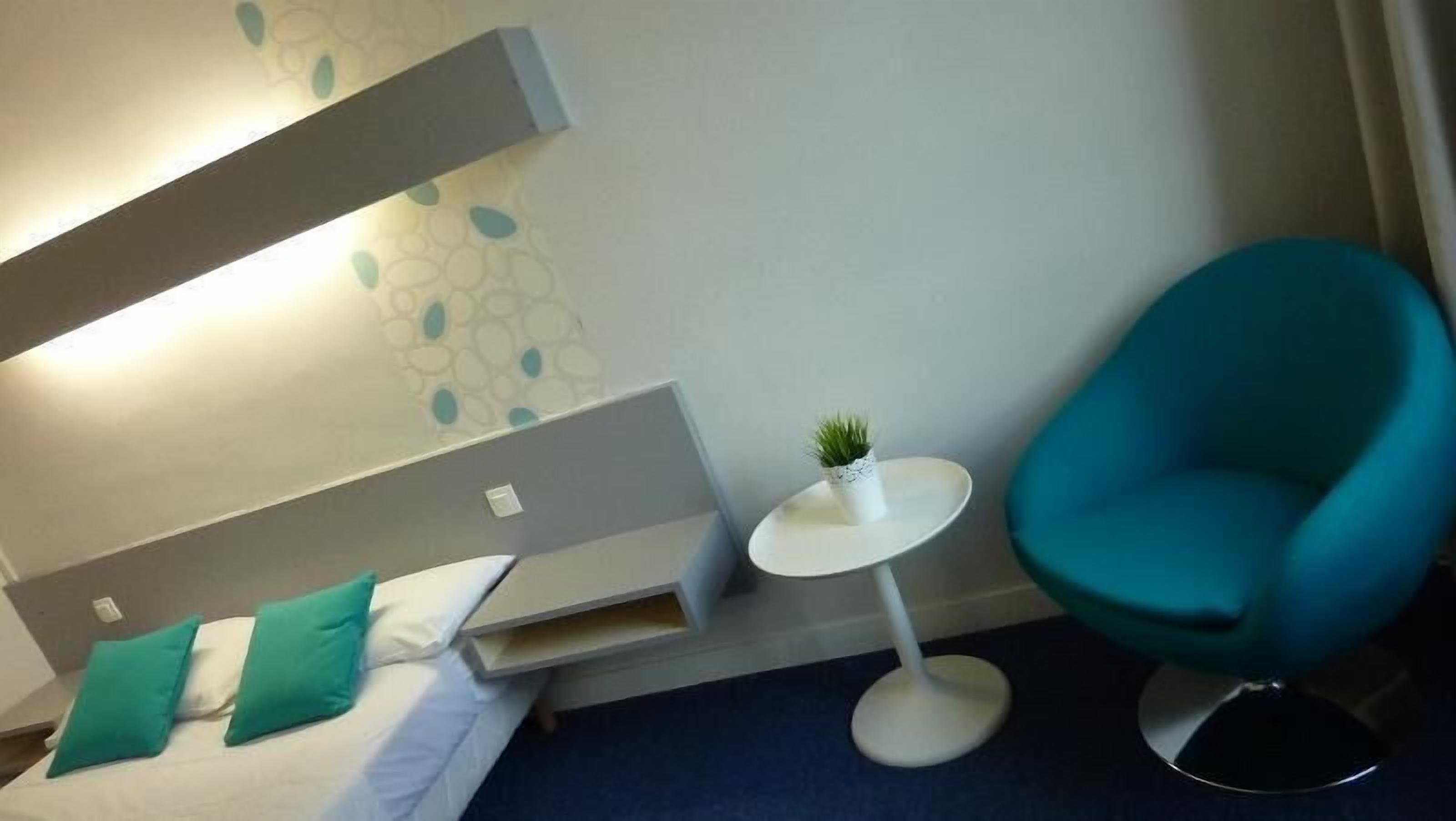 A modern hotel room with a bed, a small round table with a plant, and a teal armchair.
