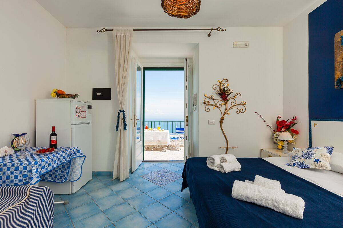 A hotel room with a bed, a desk with a table, a refrigerator, and a view of the sea through an open door.