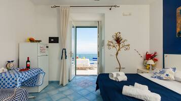 Habitación de hotel con una cama, un escritorio, un refrigerador y vista al mar a través de una puerta abierta.