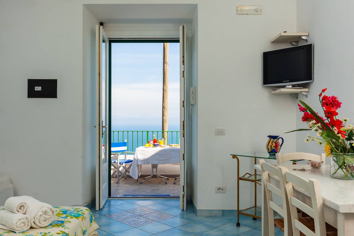 A room with a bed, a table with chairs, a TV mounted on the wall, and a view of the sea through an open door.