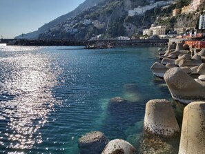 A coastal scene with clear water reflecting sunlight, rocky breakwaters, and buildings on a hillside.