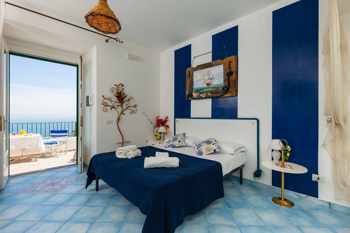 A bedroom with a bed, a bedside table, a lamp, and a view of the sea through a balcony door.