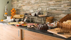 A buffet setup with various food items, including fruits, cheeses, meats, and breads, against a stone wall backdrop. - Hotel am Herkules - garni (Kassel)
