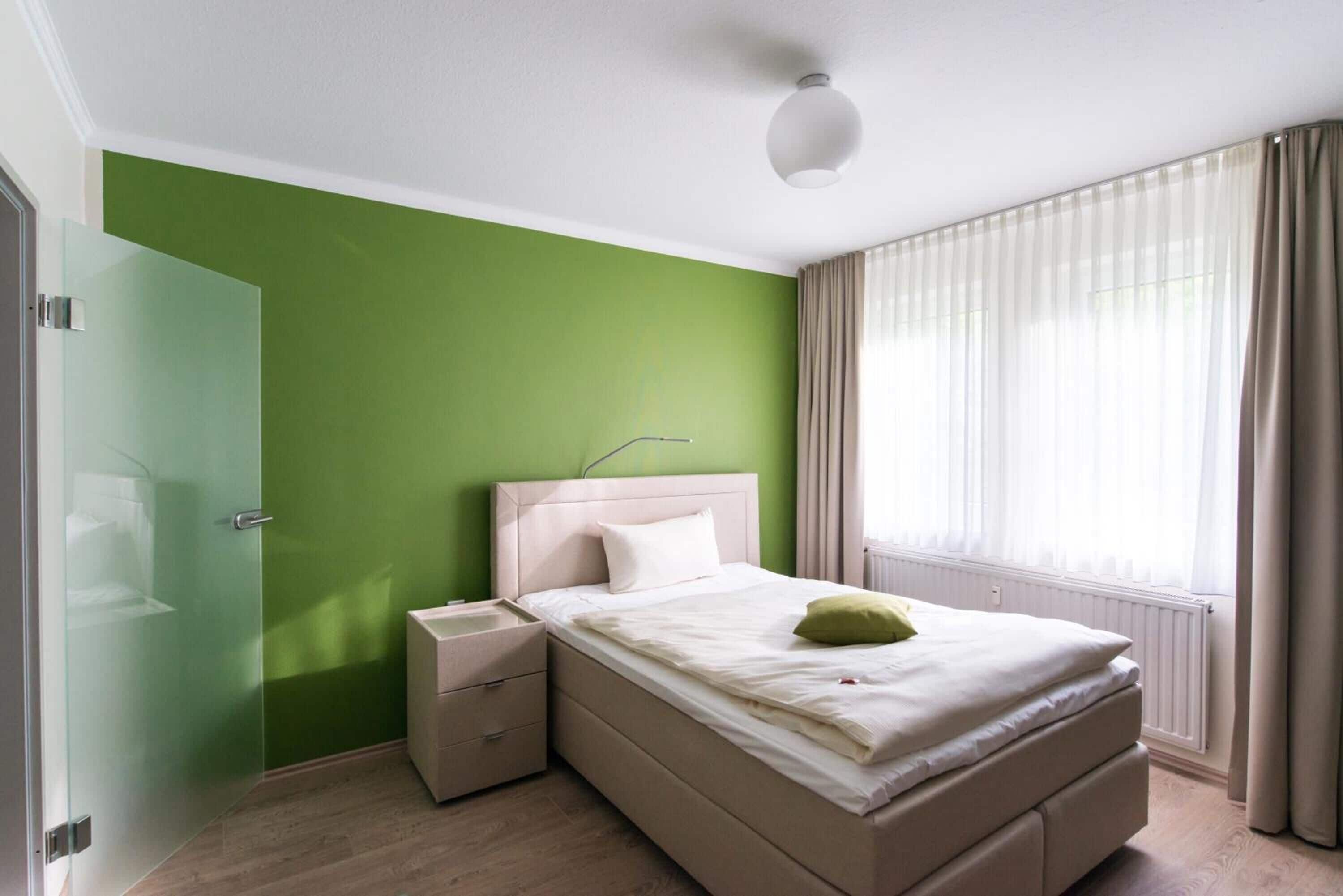 A bedroom with a green wall, a bed with a white headboard, a bedside table, and a window with curtains.