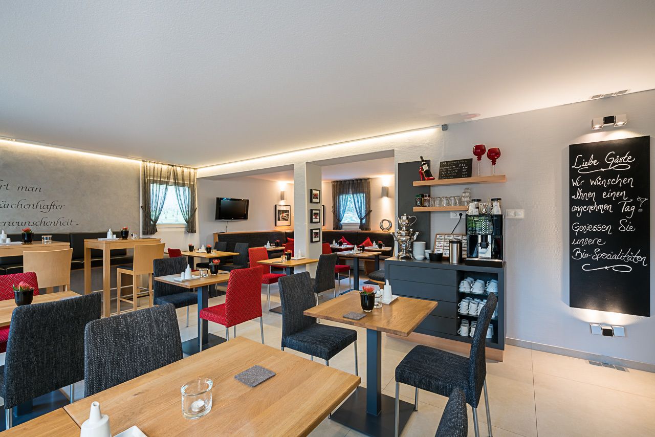 A modern restaurant with wooden tables and chairs, a chalkboard menu, and shelves with wine glasses and bottles.
