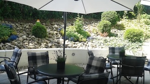 An outdoor patio with round tables and chairs, a large umbrella, and a garden with rocks and plants. - Biohotel Wilhelmshöher Tor (Kassel)