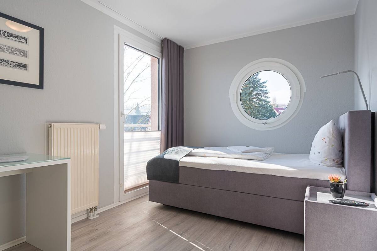 A modern bedroom with a bed, a round window, a radiator, and a desk.