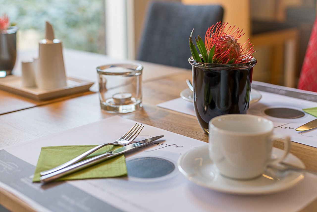 Une table à manger dressée avec une tasse blanche, une soucoupe et des couverts, le tout posé sur un set de table.