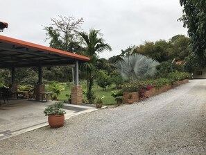 A covered outdoor seating area with tables and chairs, surrounded by a garden with various plants and a palm tree.