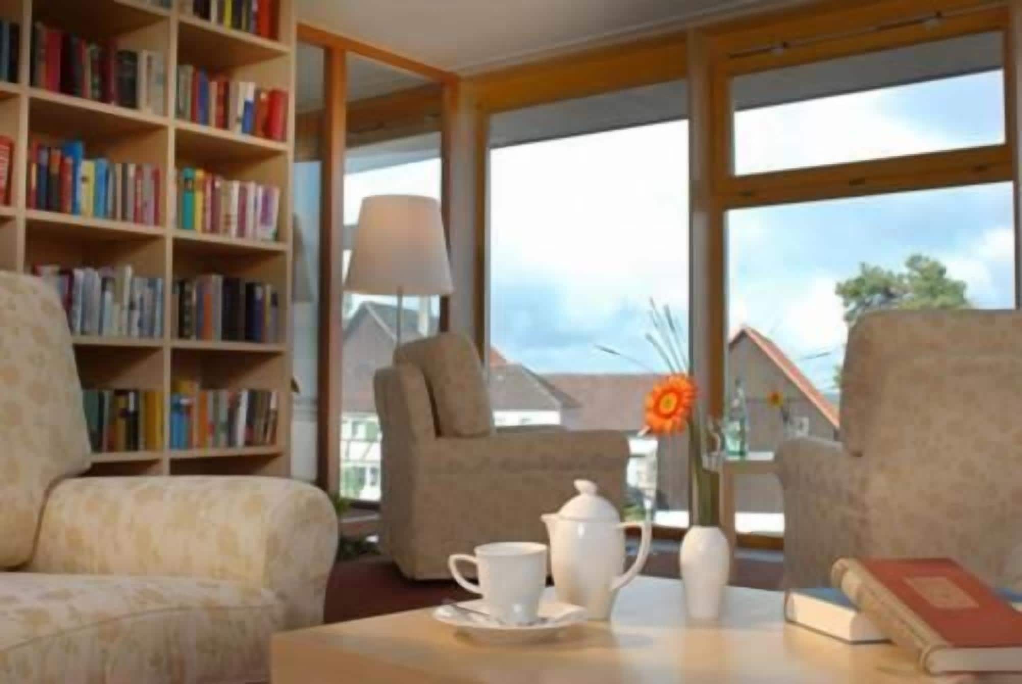 Una habitación con una estantería, dos sillones, una mesa con tetera y tazas, y un ventanal con vistas a una casa.
