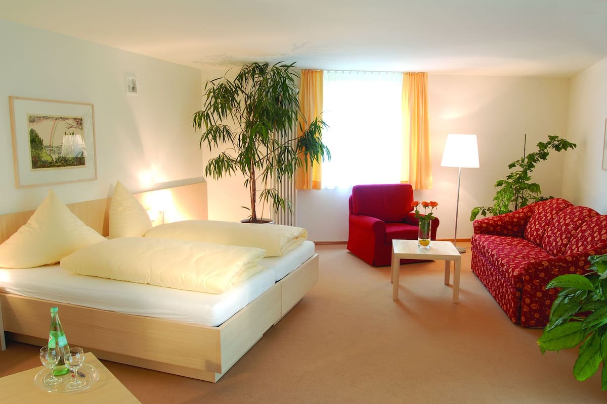 A hotel room with a bed, a red armchair, a red sofa, a small table with a lamp, and a potted plant.