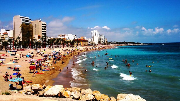 Una playa concurrida, gente nadando y tomando el sol, rodeada de edificios y palmeras.