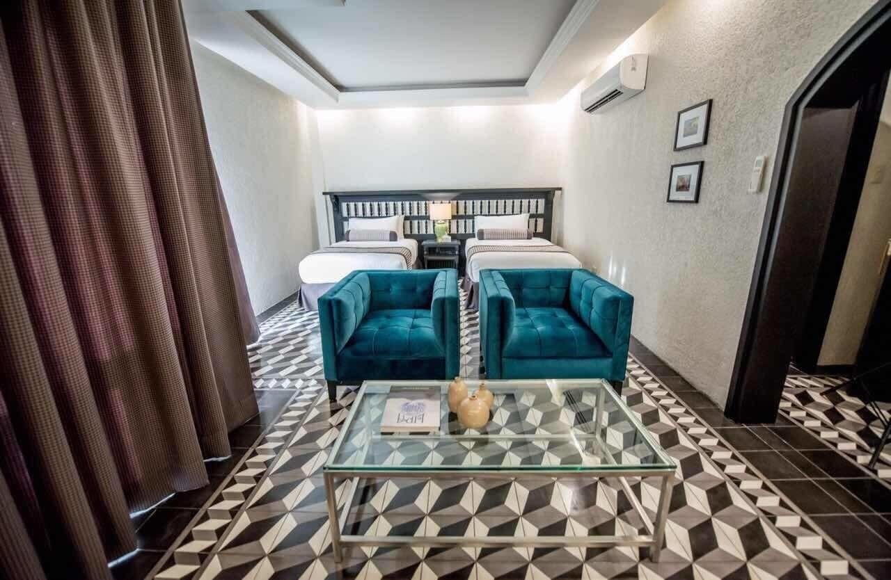 Una habitación de hotel con una cama, dos sillones azul verdoso, una mesa de centro de vidrio y un suelo con estampado geométrico.