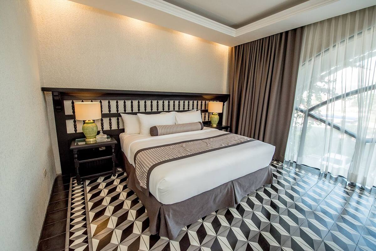A hotel room with a bed, two bedside tables, a nightstand, and a window with curtains.