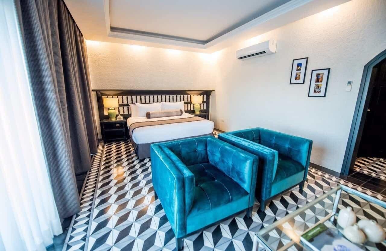 Une chambre d’hôtel avec un lit, deux fauteuils bleus, une table de chevet et un sol carrelé.