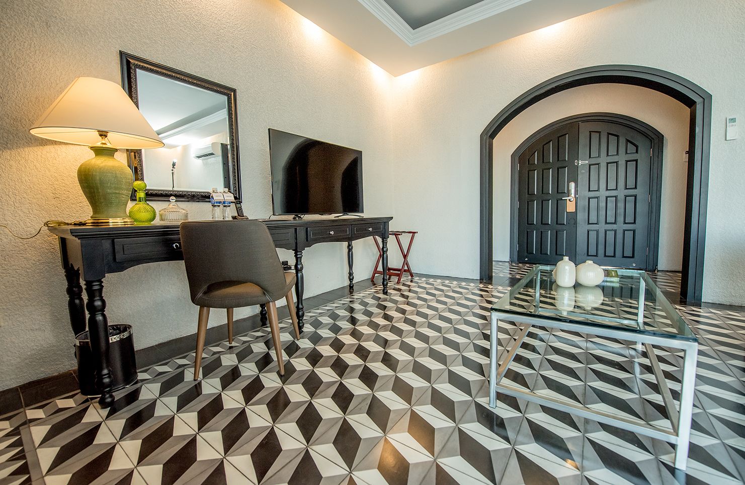 A hotel lobby with a geometric patterned floor, a desk with a lamp, a chair, a television, and a glass table with decorative items.