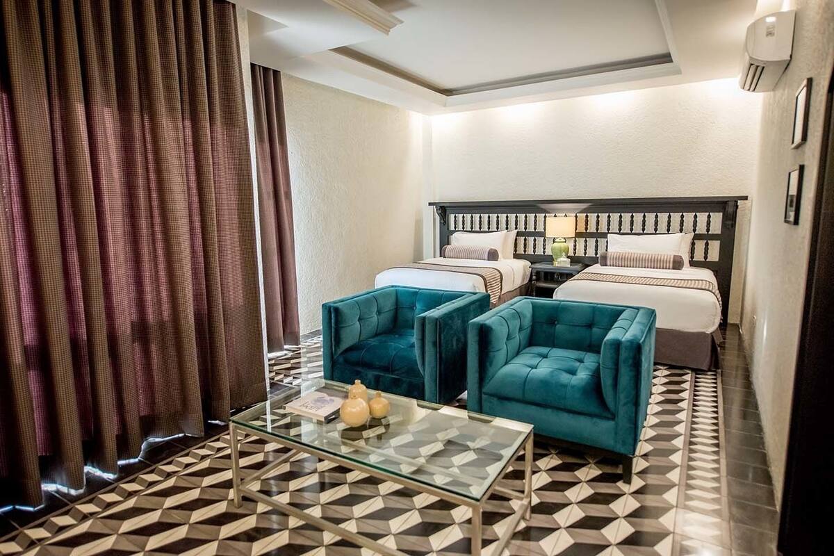A hotel room with a bed, two blue armchairs, a glass coffee table, and a patterned floor.