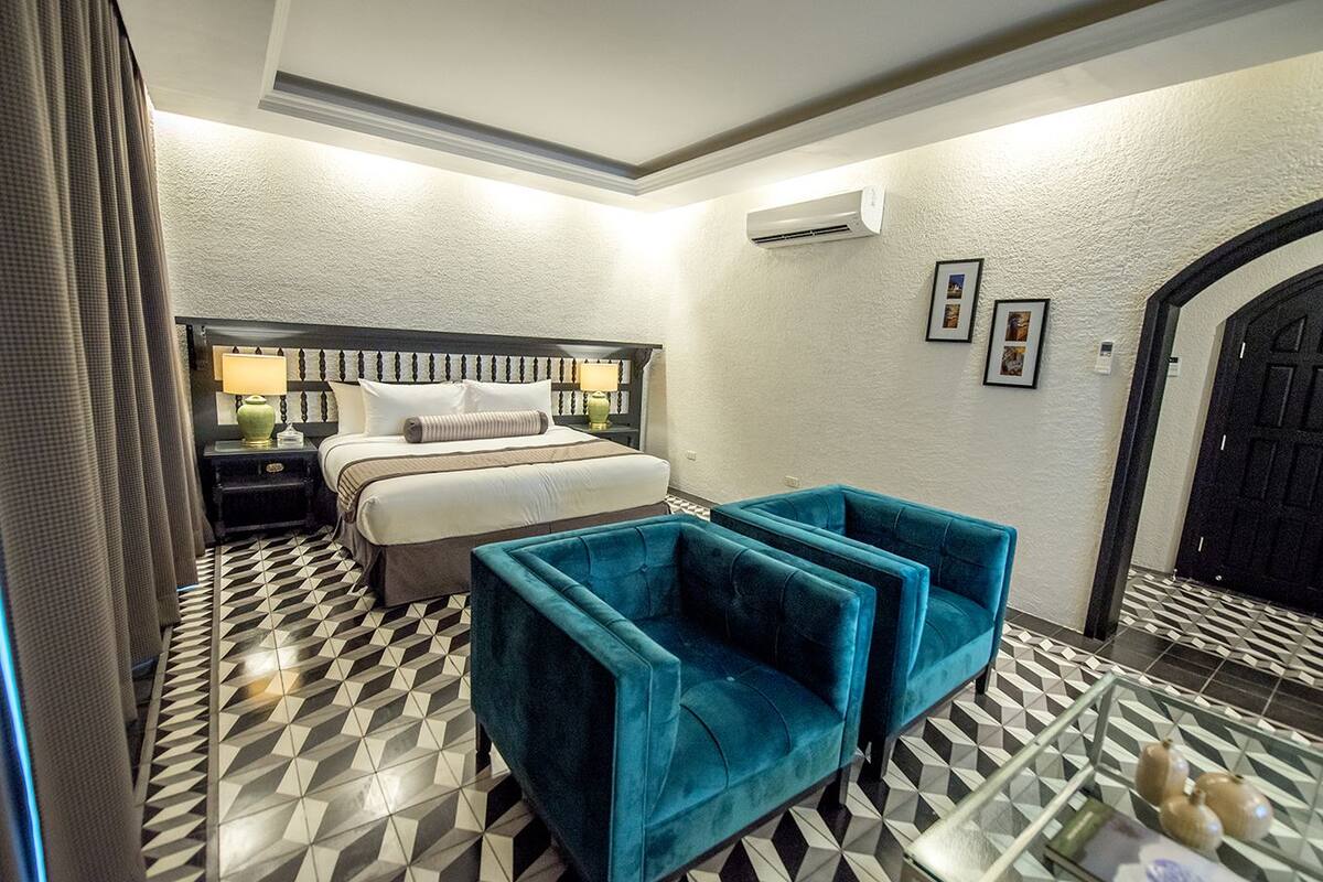 A hotel room with a bed, two bedside tables, a lamp, a blue armchair, and a geometric patterned floor.