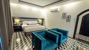 Una habitación de hotel con cama, dos mesitas de noche, una lámpara, un sillón azul y un suelo con estampado geométrico.