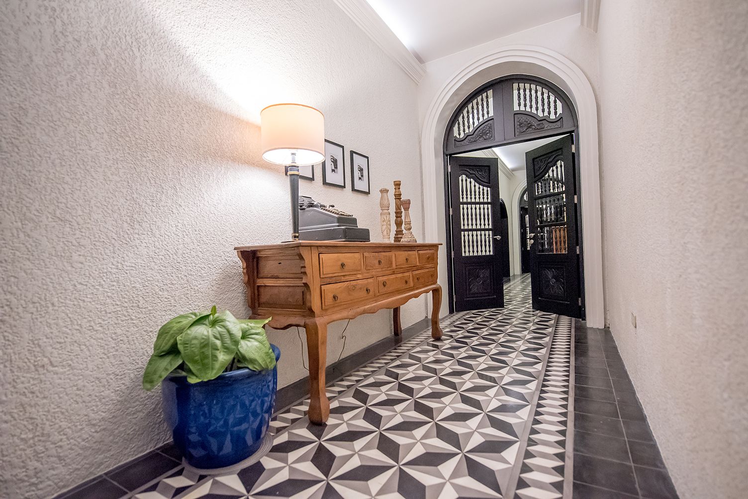 A hallway with a geometric patterned floor, a wooden dresser, a wall-mounted lamp, and a potted plant.