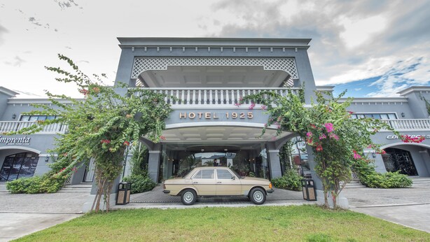 A hotel entrance with a vintage car parked in front, surrounded by greenery and a clear sky.