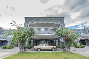 Een hotelentree met een oldtimer ervoor, omgeven door groen en een heldere lucht.