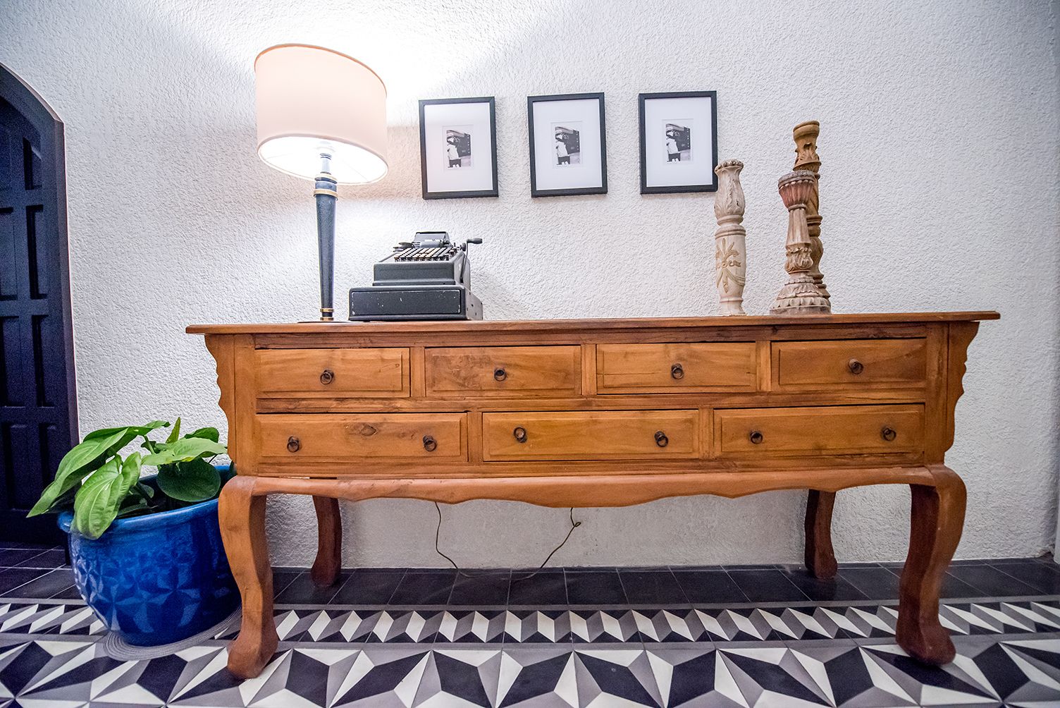 A wooden dresser with drawers, a lamp, and framed pictures on a textured wall.