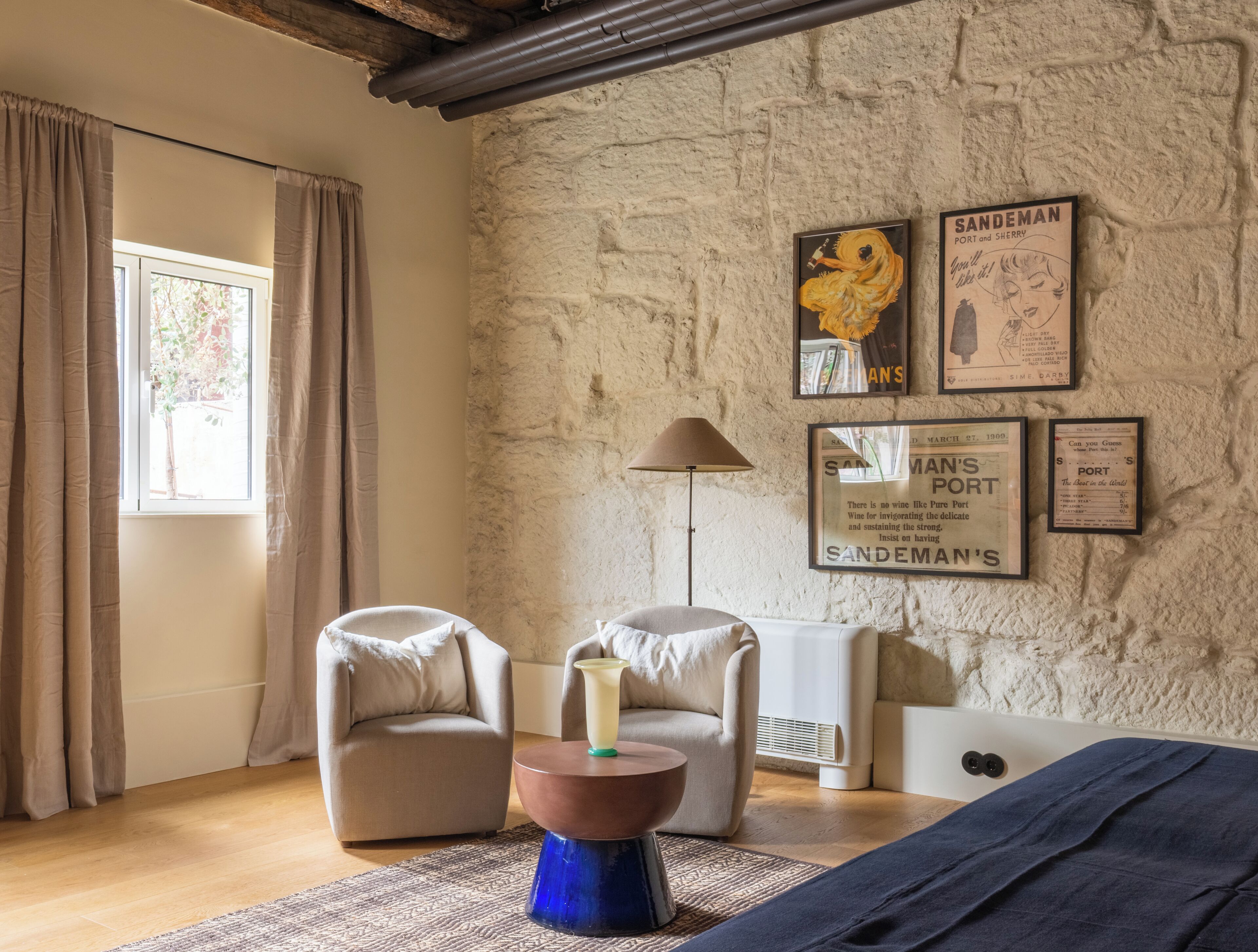 A room with a stone wall, two armchairs, a small table, a lamp, and framed posters.