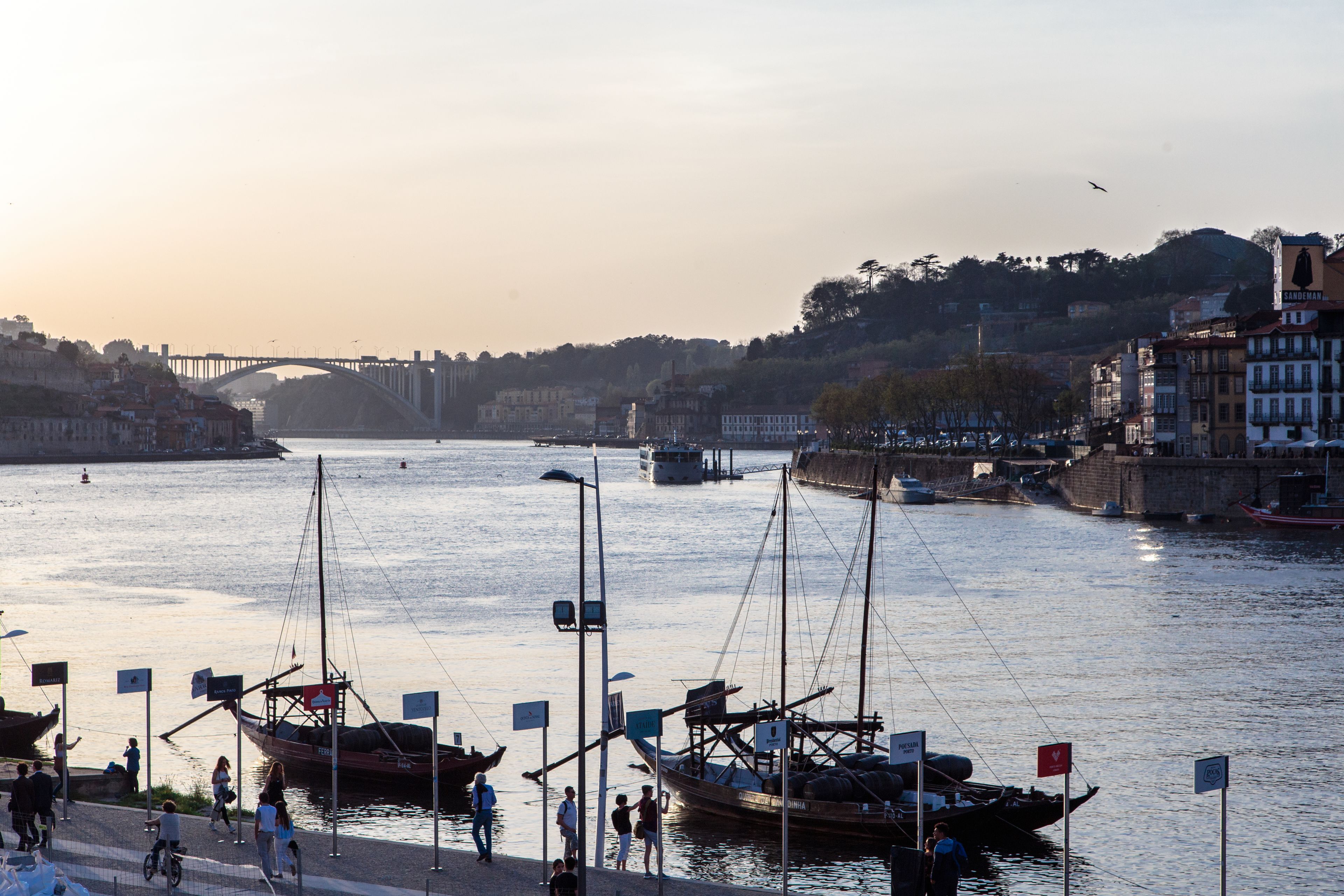 A riverside view with boats docked, a bridge in the background, and buildings along the riverbank.