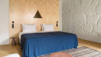 A bedroom with a wooden headboard, a blue bedspread, a patterned rug, and a small round ottoman.