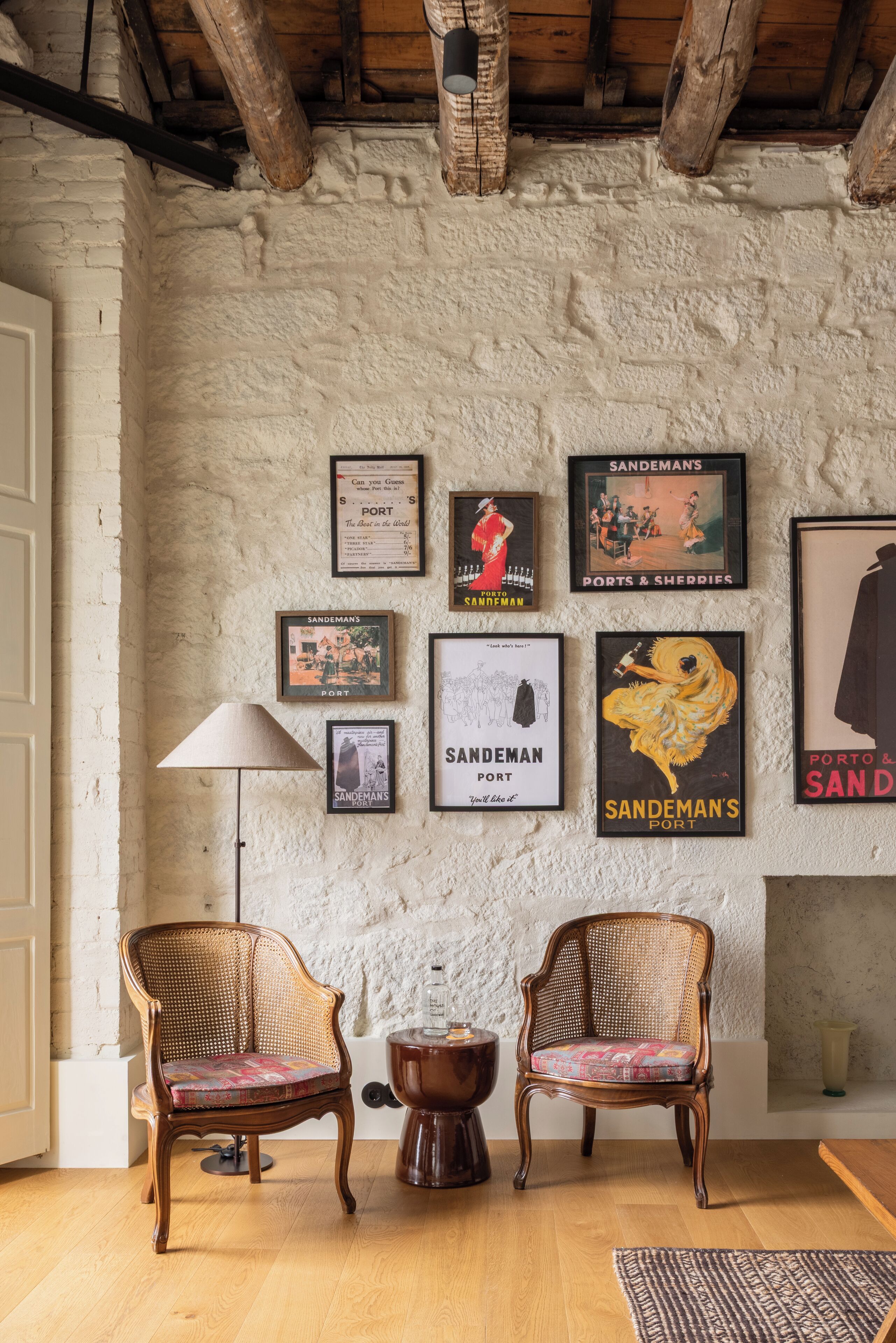 Ein Zimmer mit rustikalen Holzbalken, einer weißen, strukturierten Wand und einer Sammlung gerahmter Vintage-Poster. Zwei Korbstühle mit bunten Kissen und ein kleiner Holztisch sind ebenfalls vorhanden.