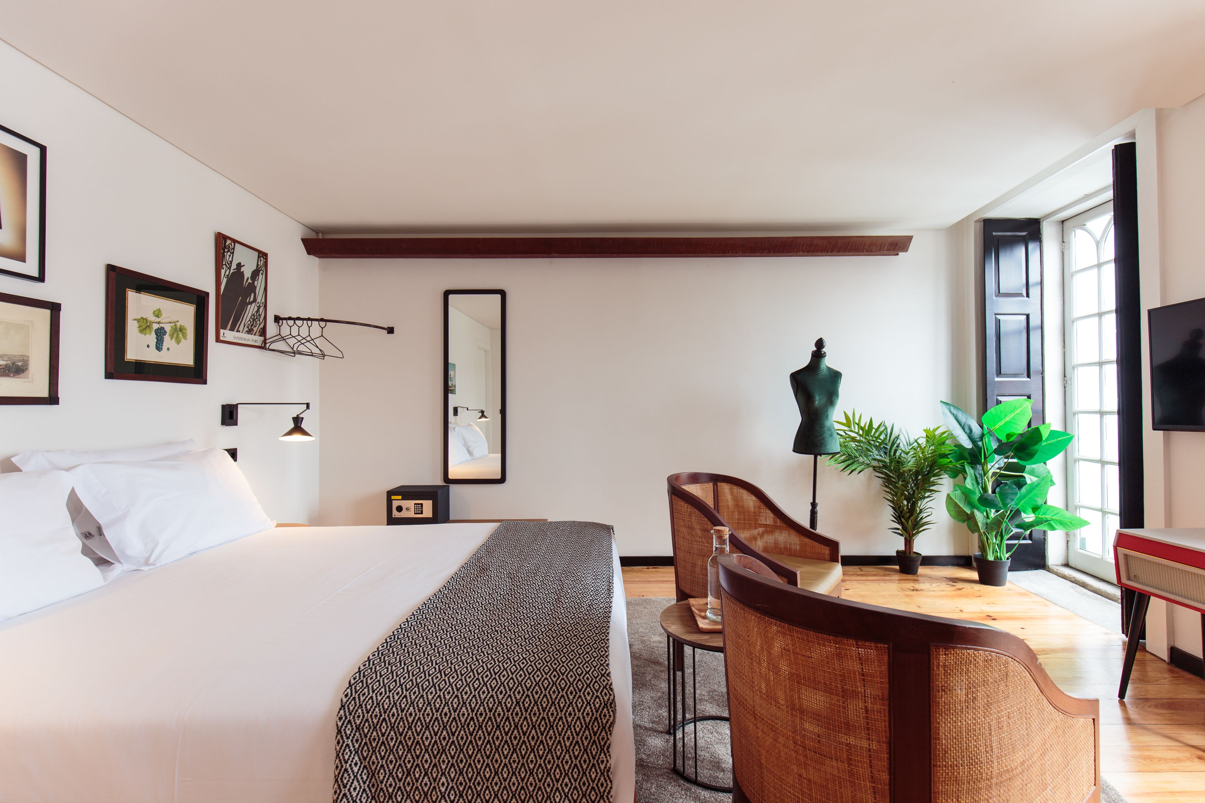 A hotel room with a bed, a desk with a chair, a potted plant, a mirror, and framed pictures on the wall.