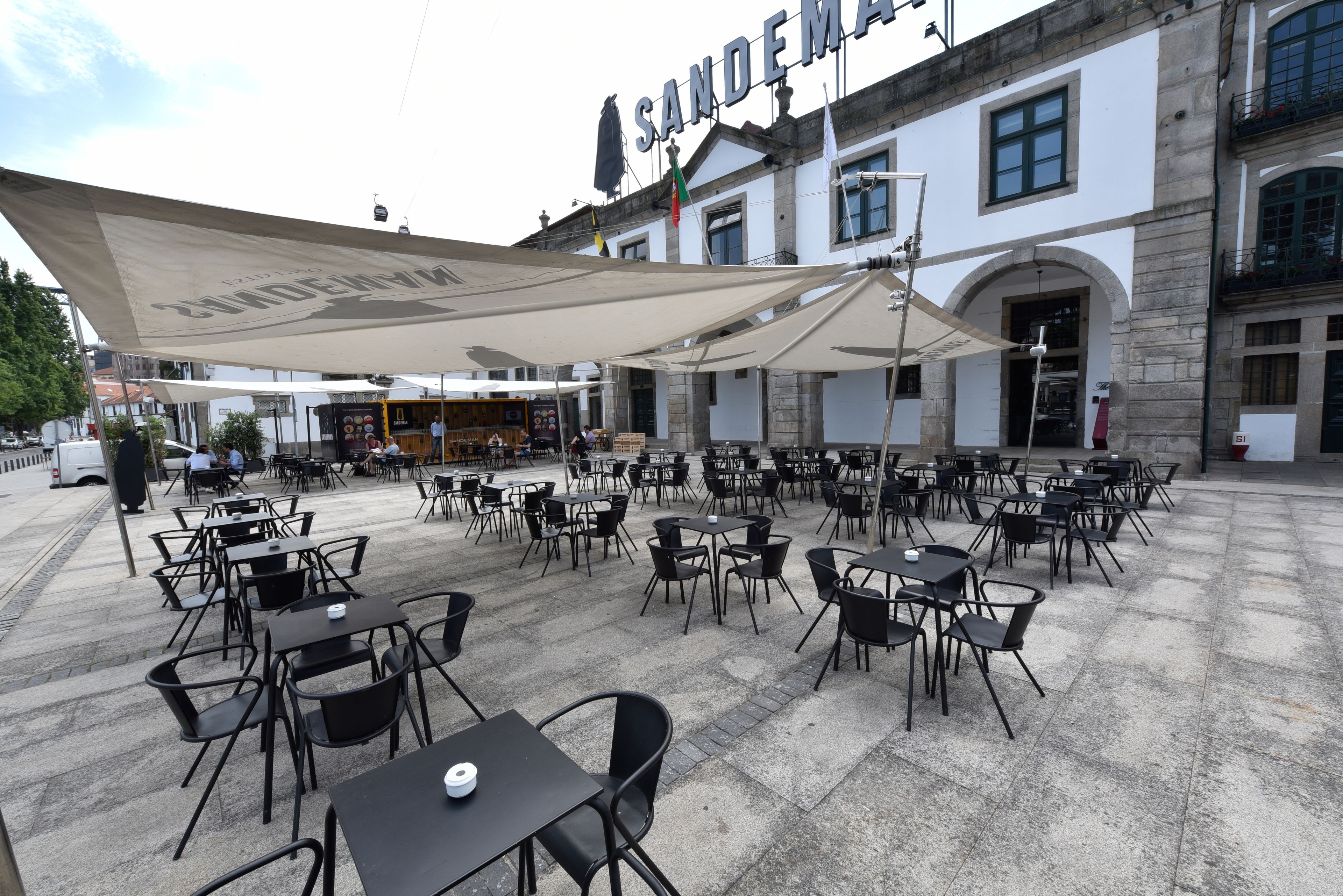 An outdoor cafe area with multiple tables and chairs, a large canopy, and a building with the name "SANDEMA" in the background.