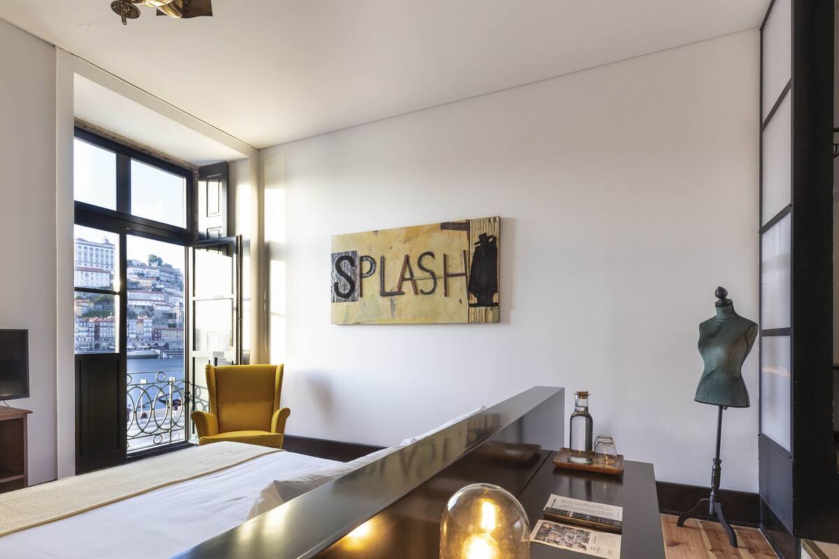 Une chambre d’hôtel moderne dotée d’une grande fenêtre, d’un fauteuil jaune, d’un téléviseur à écran plat, d’une table d’appoint en bois et d’une œuvre d’art murale sur laquelle figure le mot « SPLASH ».