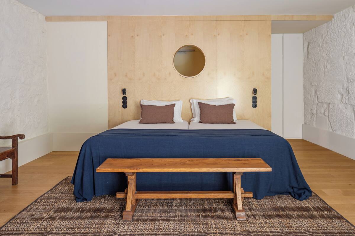 Une chambre avec une tête de lit en bois, un miroir rond, deux lits avec des oreillers marron, un banc en bois et un tapis à motifs.