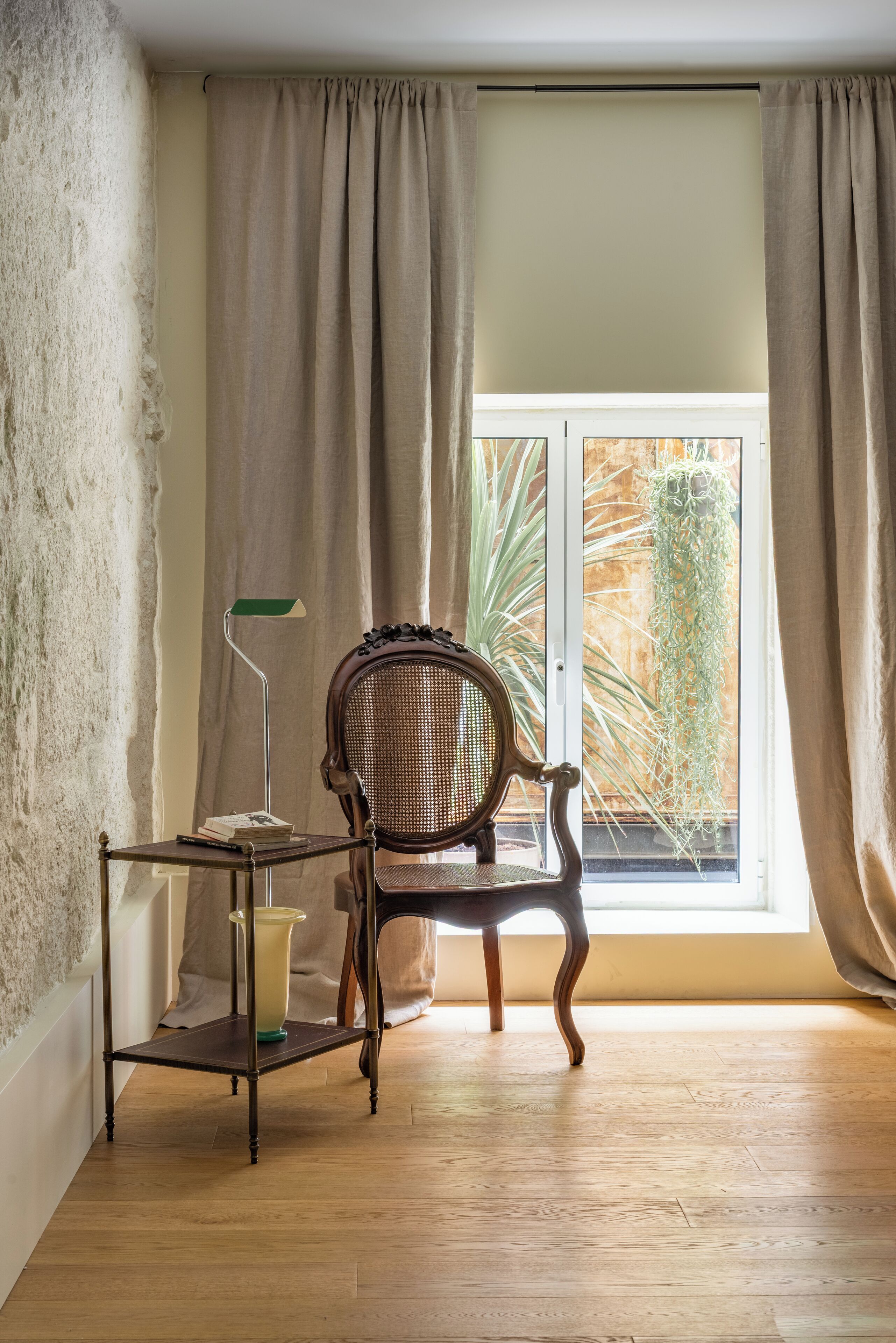 Une pièce dotée d'un plancher en bois, d'une chaise en osier, d'une petite table avec une lampe, et offrant une vue sur la verdure à travers une fenêtre ornée de rideaux beiges.