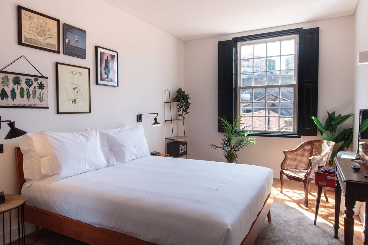 Une chambre bien rangée, avec un lit, une table de chevet, une lampe, une chaise et une fenêtre donnant sur des immeubles.