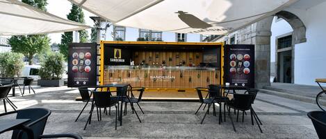 An outdoor seating area with tables and chairs under a canopy, a bar with a sign reading "Sandeman," and a stone building in the background.