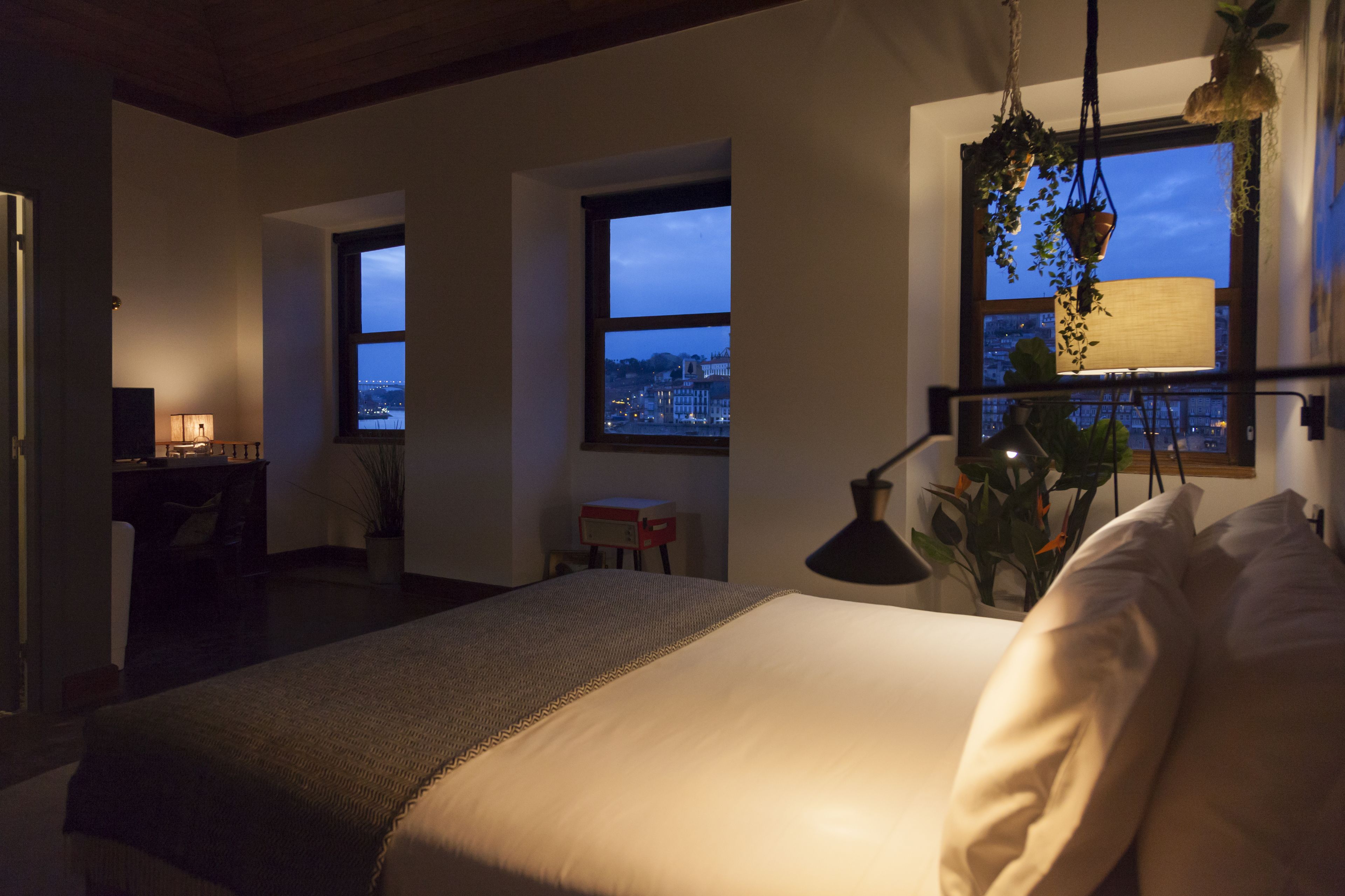 Un dormitorio con una cama grande, un escritorio con lámpara y vistas a la ciudad a través de dos ventanas.