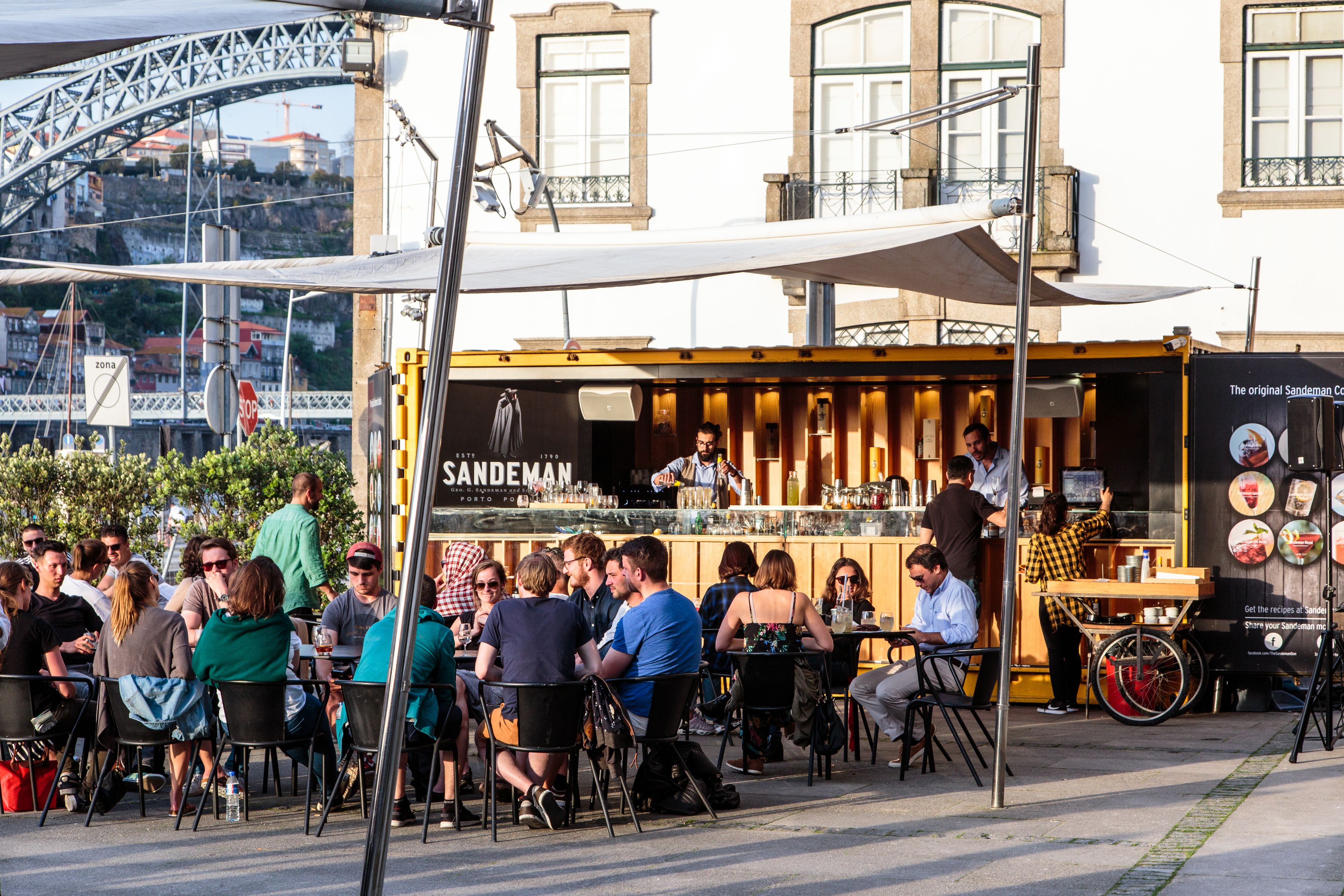 A street-side café with outdoor seating, a visible menu board, and a view of a bridge in the background.