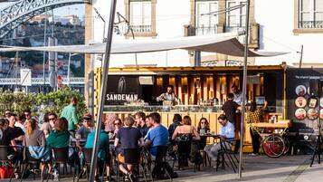 Um café à beira da rua com mesas ao ar livre, um quadro de ementa visível e vista para uma ponte ao fundo.