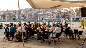 Un área de comedor al aire libre con mesas y sillas bajo un toldo grande, con vista a un frente marítimo y edificios al fondo.