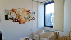 A modern living room with a white sofa set, a small white table, and a wooden dining table. There are abstract paintings on the wall and a window with a view of buildings. - Residence Nizza - Torino (Turin)