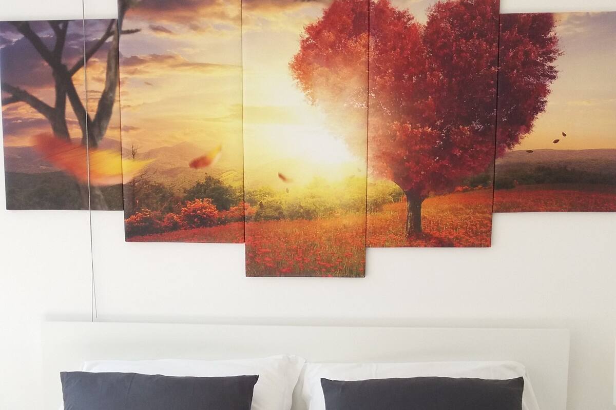 A bed with white bedding and two dark pillows, a white headboard, and a wall-mounted multi-panel artwork depicting a tree and landscape.