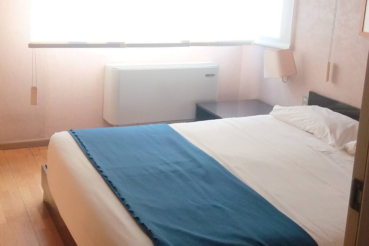 A bedroom with a bed, a nightstand, a window with blinds, a radiator, and a wall-mounted lamp.