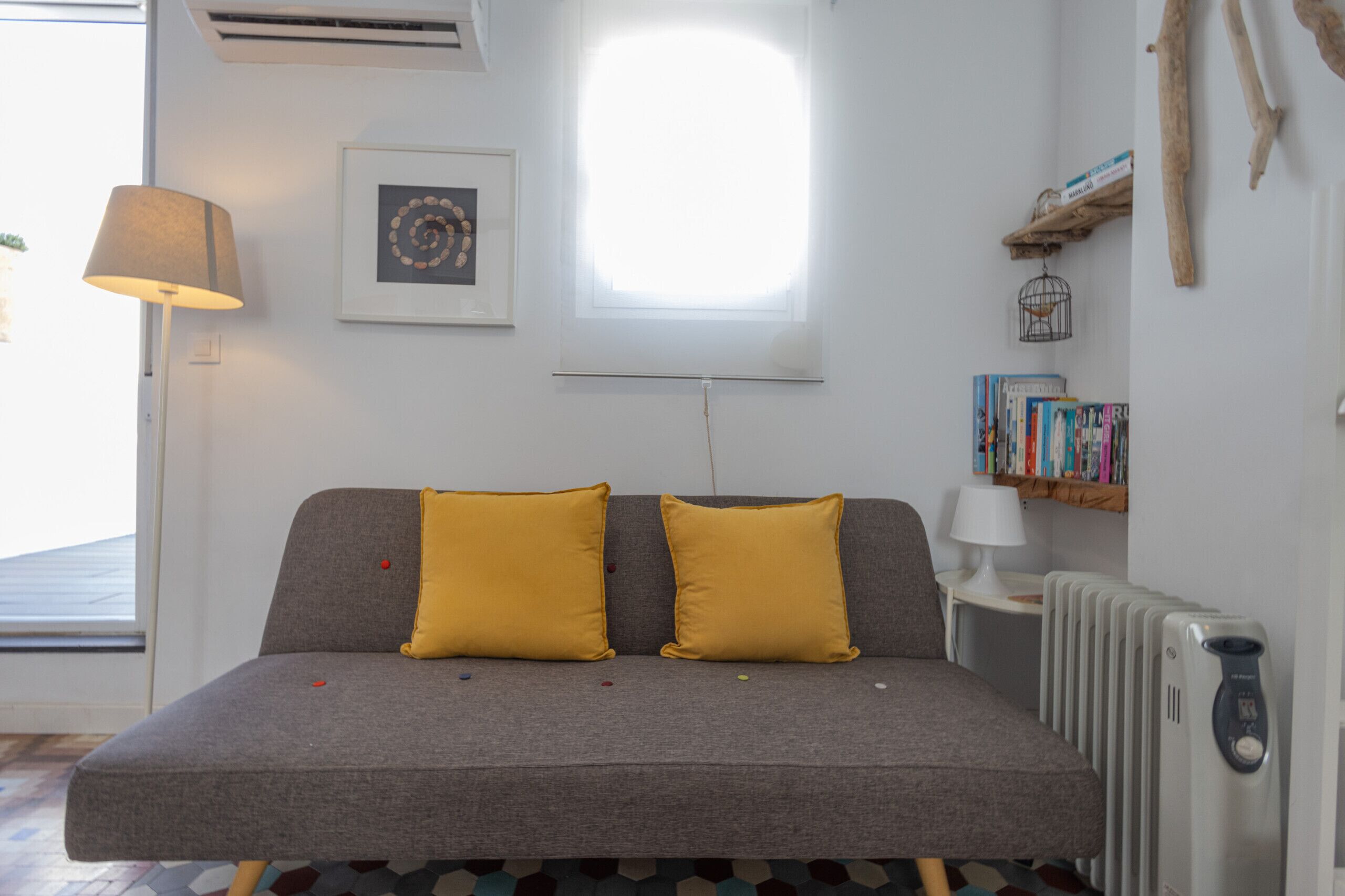 Ein modernes Wohnzimmer mit einem grauen Sofa, zwei gelben Kissen, einem Beistelltisch mit Lampe, einem Bücherregal voller Bücher, einer Heizung und einem gerahmten Bild an der Wand.