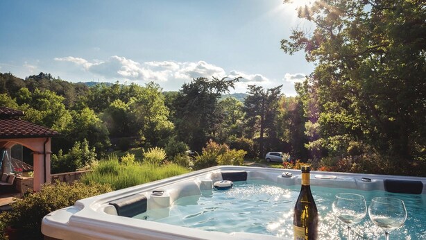 Un jacuzzi con una botella de vino y copas, rodeado de exuberante follaje y un pabellón.