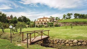 Un puente de piedra sobre un arroyo que conduce a una casa grande con jardín y viñedo al fondo.