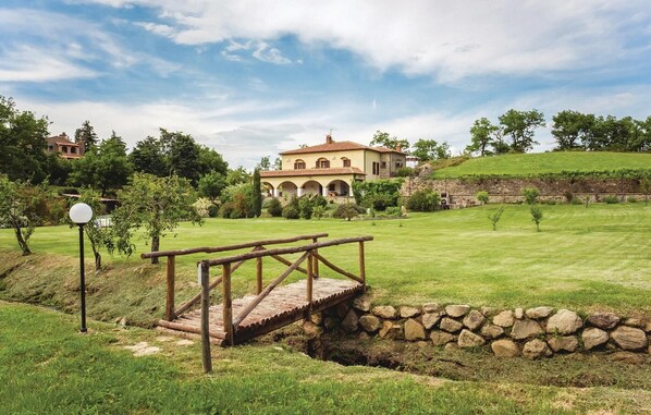 Un pont de pierre enjambe un ruisseau et mène à une grande maison, avec un jardin et un vignoble en arrière-plan.