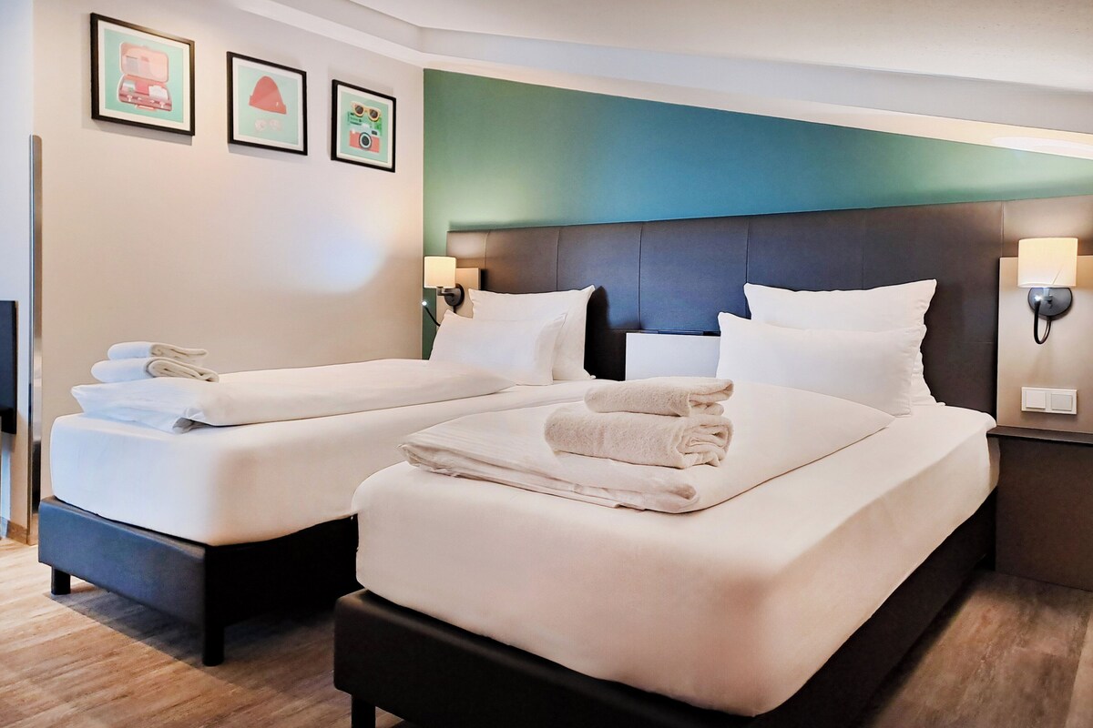 A hotel room with two beds, a teal accent wall, and wall-mounted lamps.